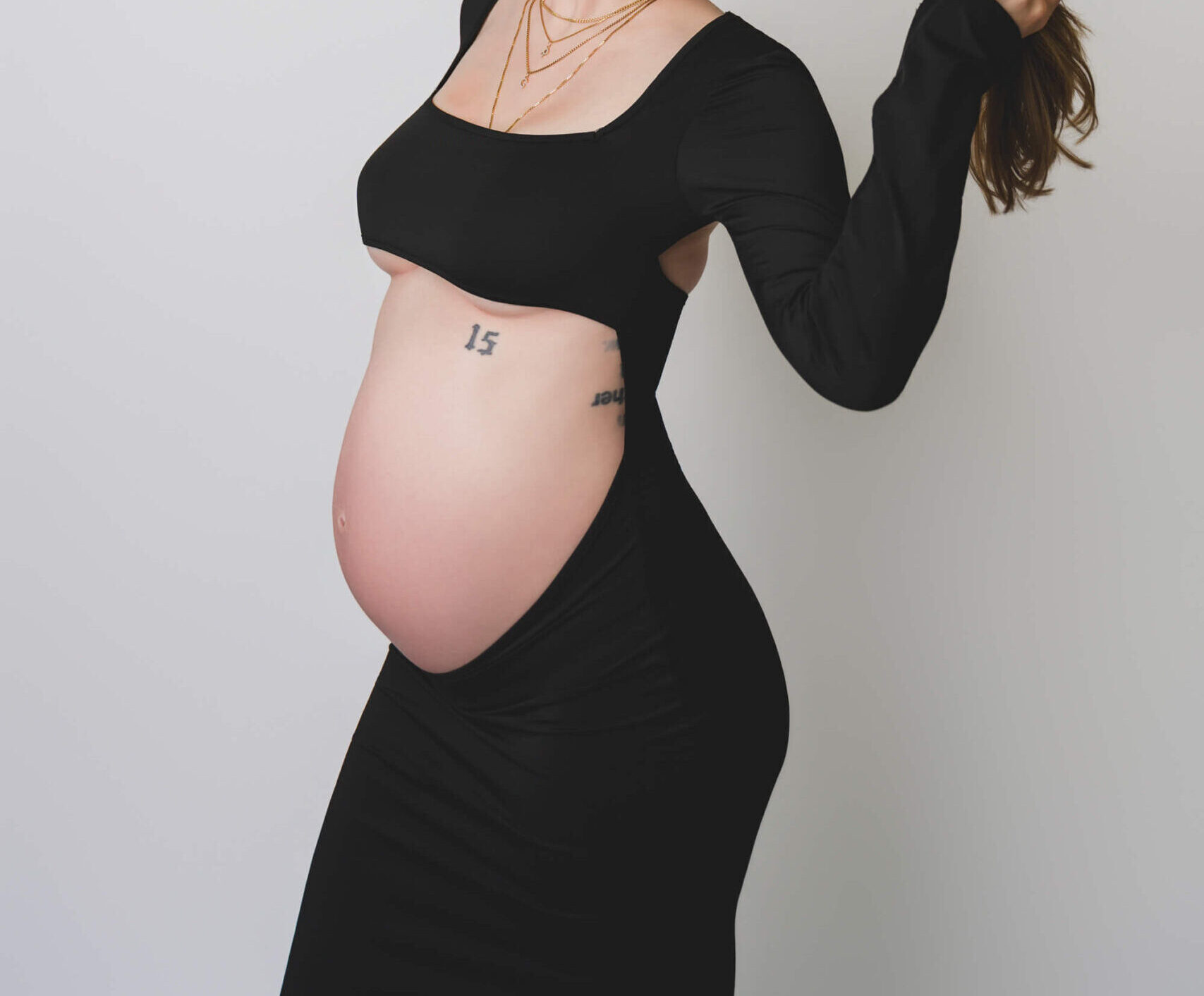 A mother to be twirls her hair while standing in a studio in a black maternity dress with the bump out