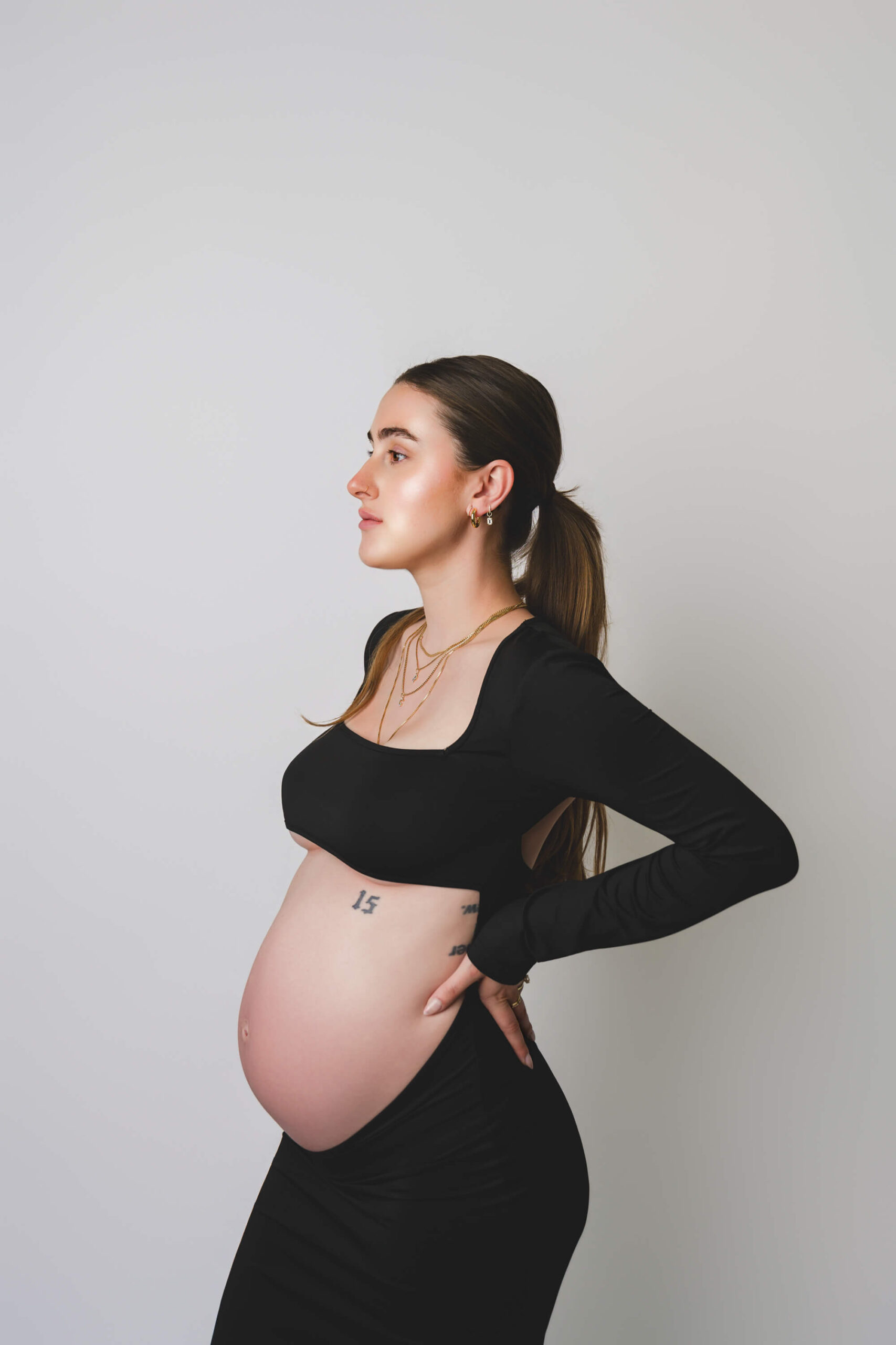 A mom to be in a black open belly bump rests her hands on her back while standing in a white studio after visiting baby shower venues in san francisco