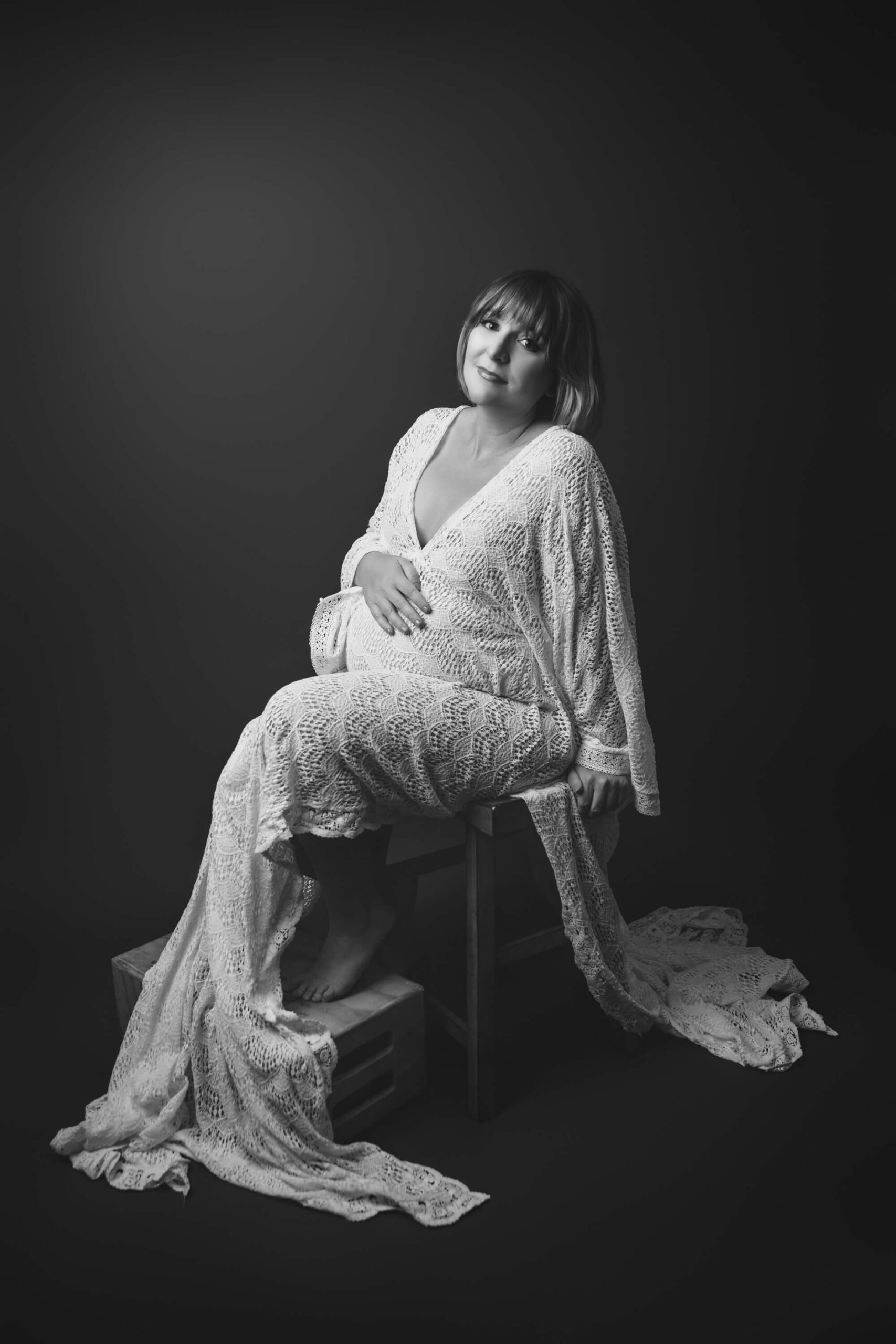 A pregnant woman sits on a stool in a lace maternity gown with a hand on her bump in black and white after visiting birth centers in san francisco