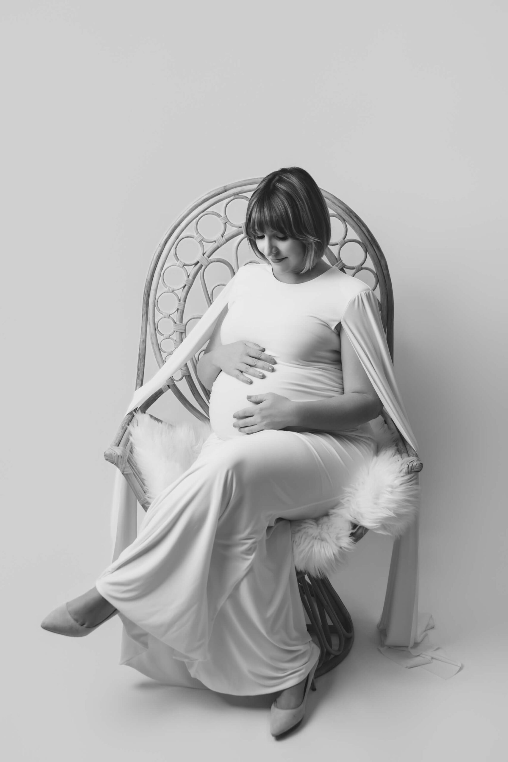 A smiling pregnant woman sits in a wicker hair in a white maternity gown smiling down at her hands on her bump after visiting birth centers in san francisco