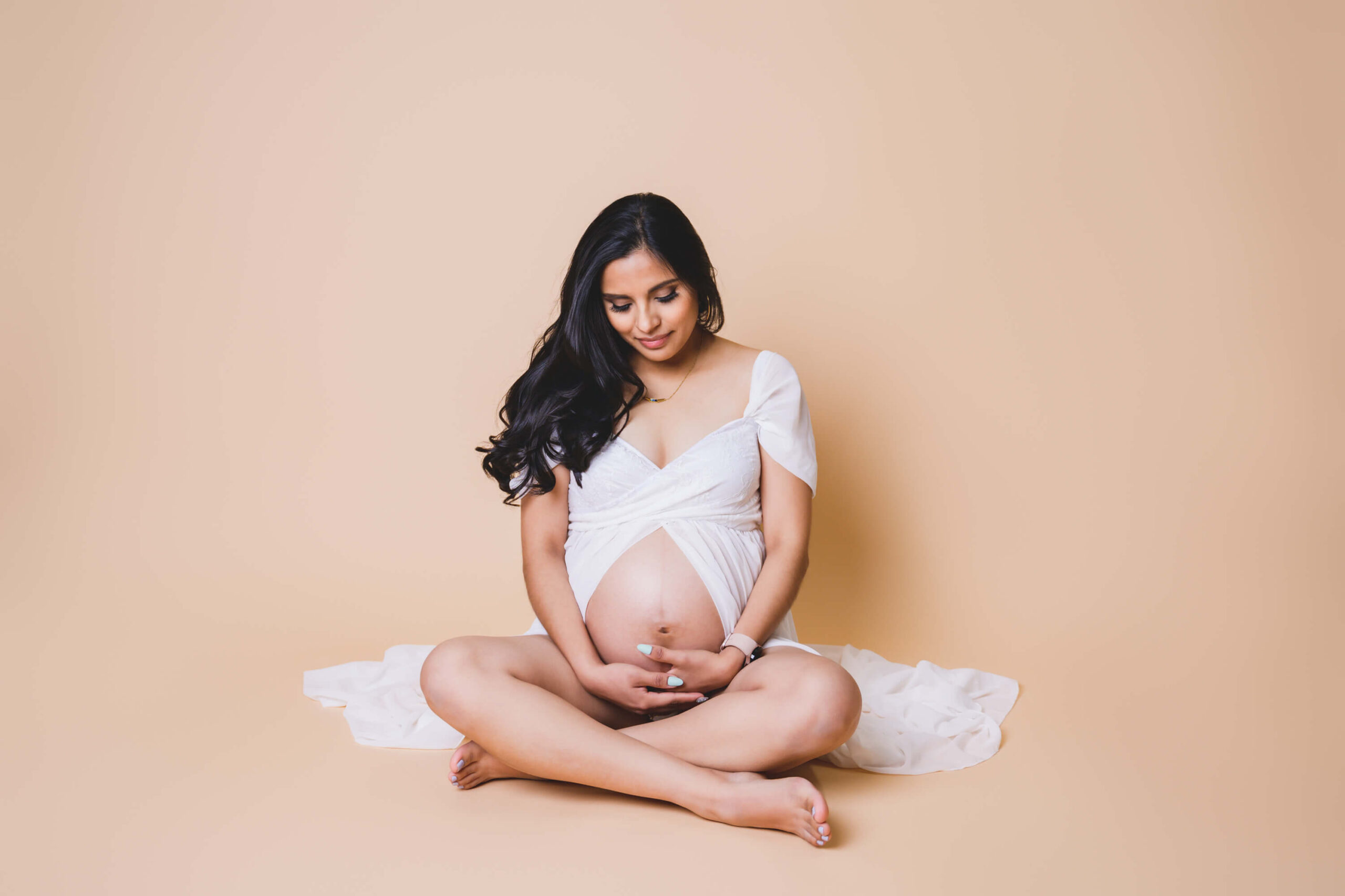 A pregnant woman smiles down to her bump in her hands while sitting on the studio floor in an open front white maternity gown