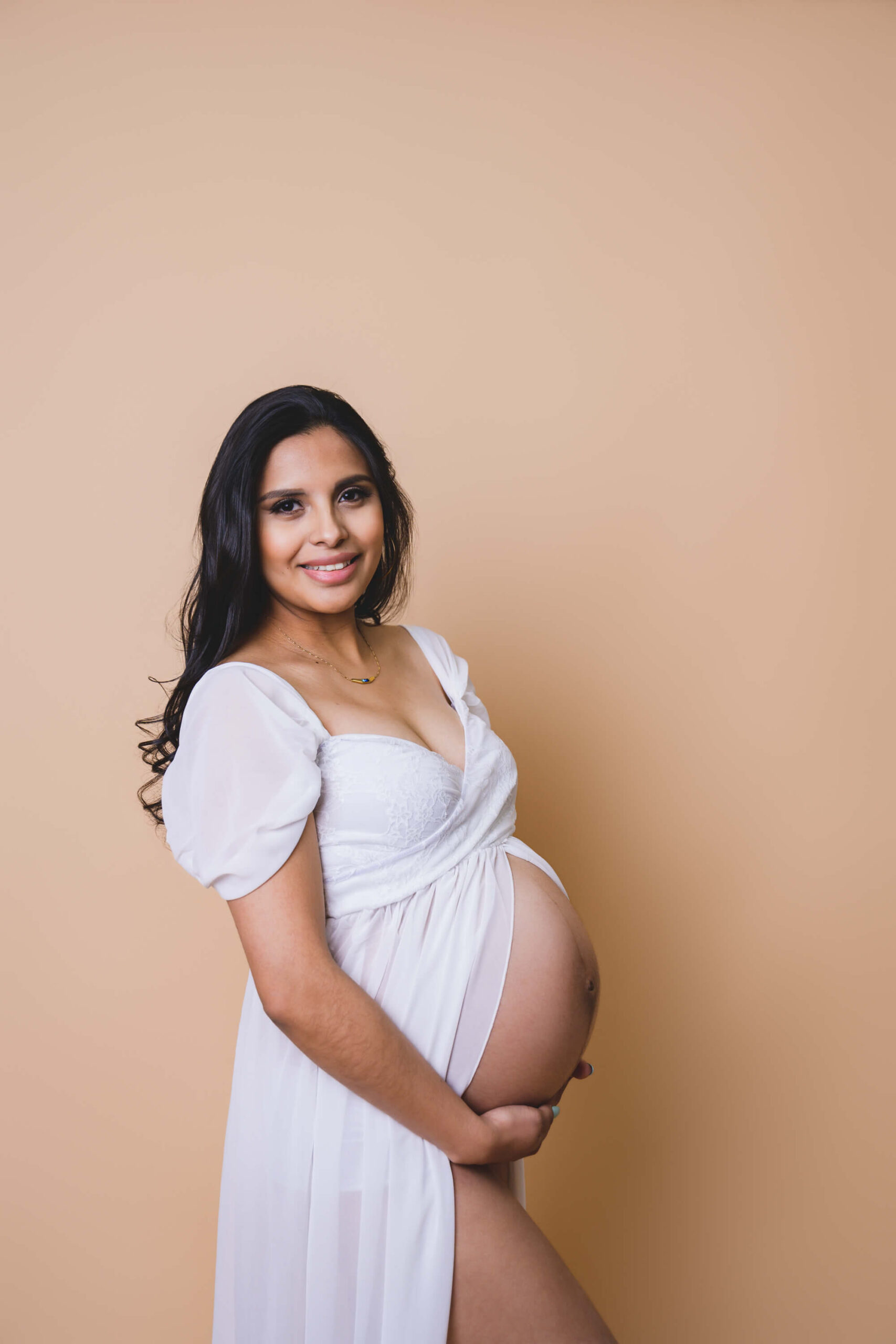 A smiling mother to be in an open front white maternity gown holding hands under her bump in a studio after meeting doulas in marin