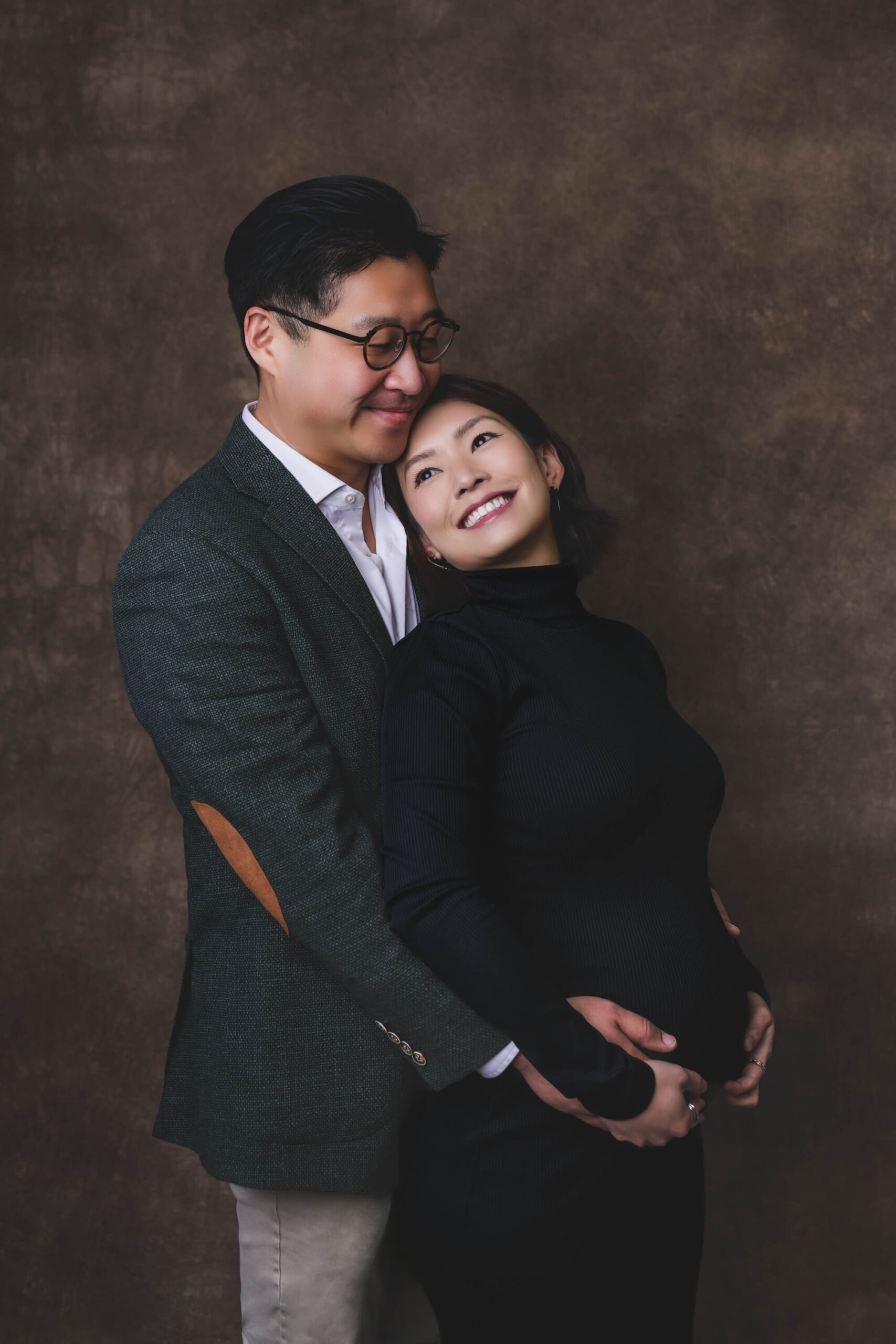 A smiling woman in a black gown leans into the chest of her husband in a green sports coat as they both hold the bump