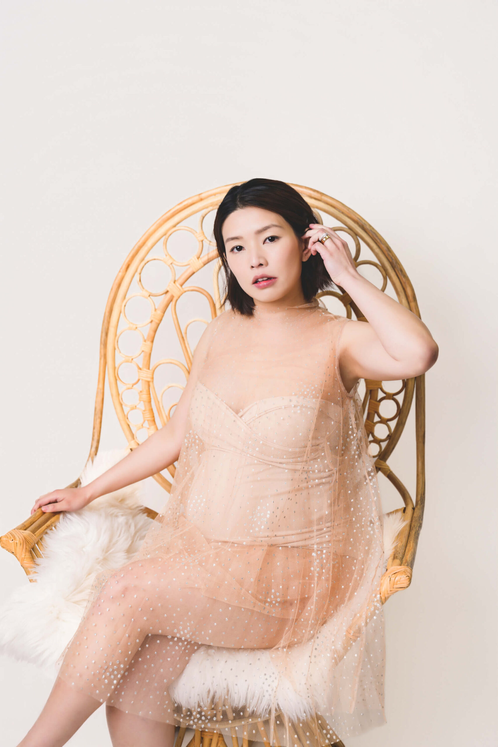 A pregnant woman in a sheer pink fabric and nude bodysuit sits in a wicker hair pulling hair behind her ear after meeting midwives in san francisco