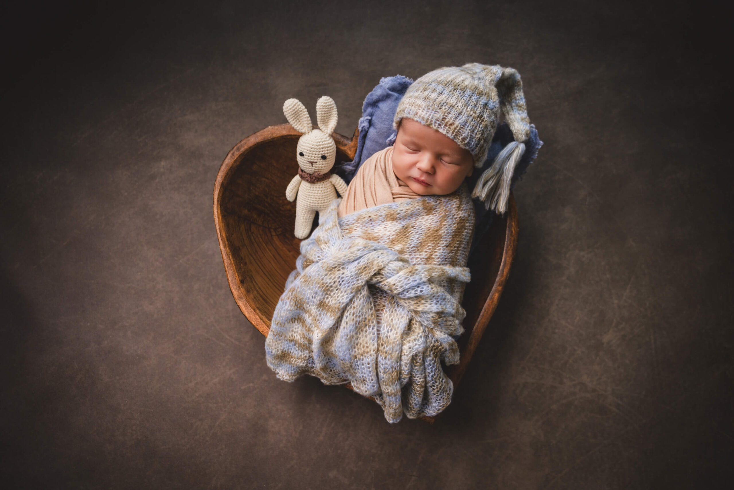 A newborn baby sleeps in a blue knit blanket and matching sleep cap in a wooden bowl with a tiny stuffed bunny in a studio
