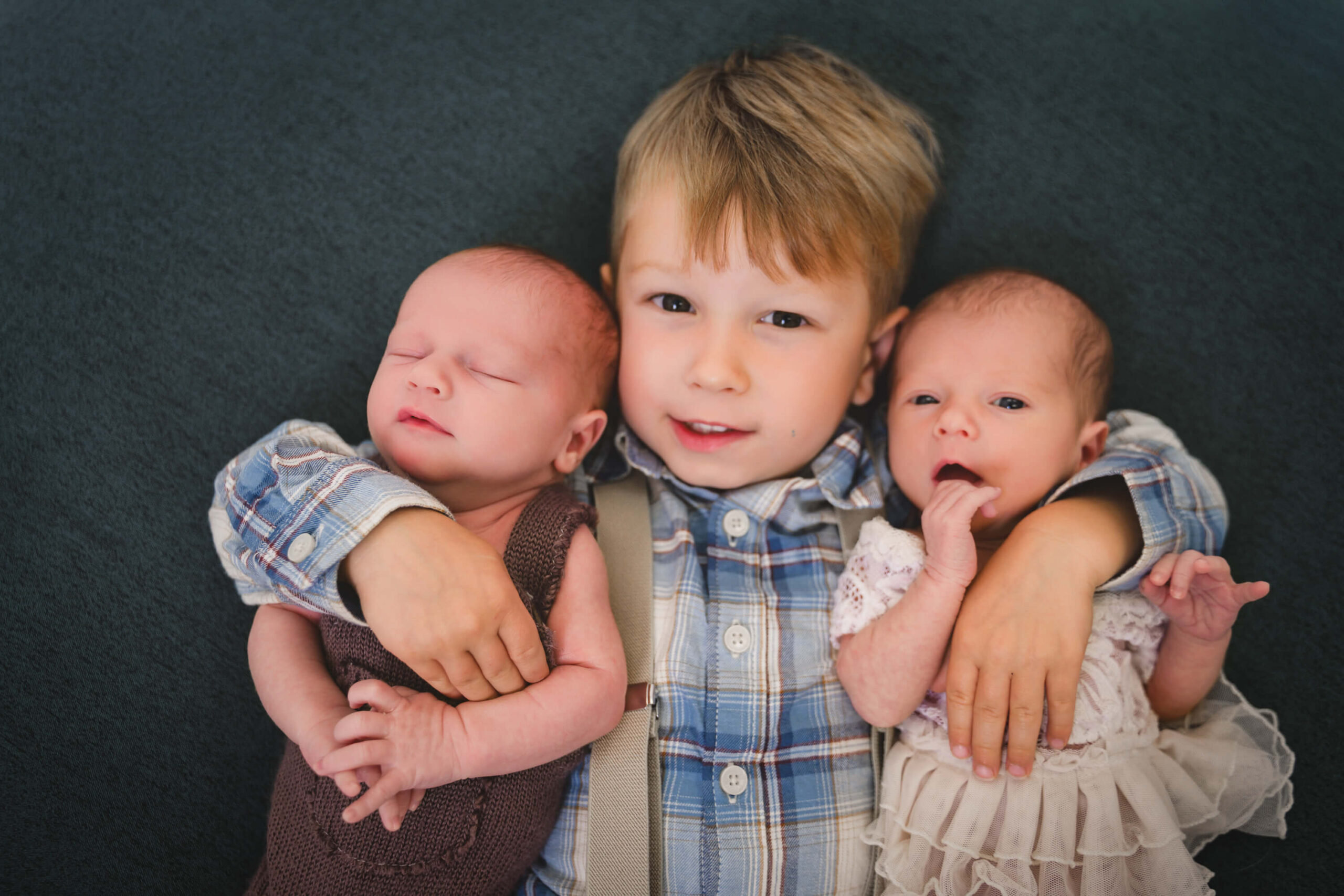 A toddler boy in a blue plaid shirt lays with arms around his newborn twin brother and sister on a blue bed
