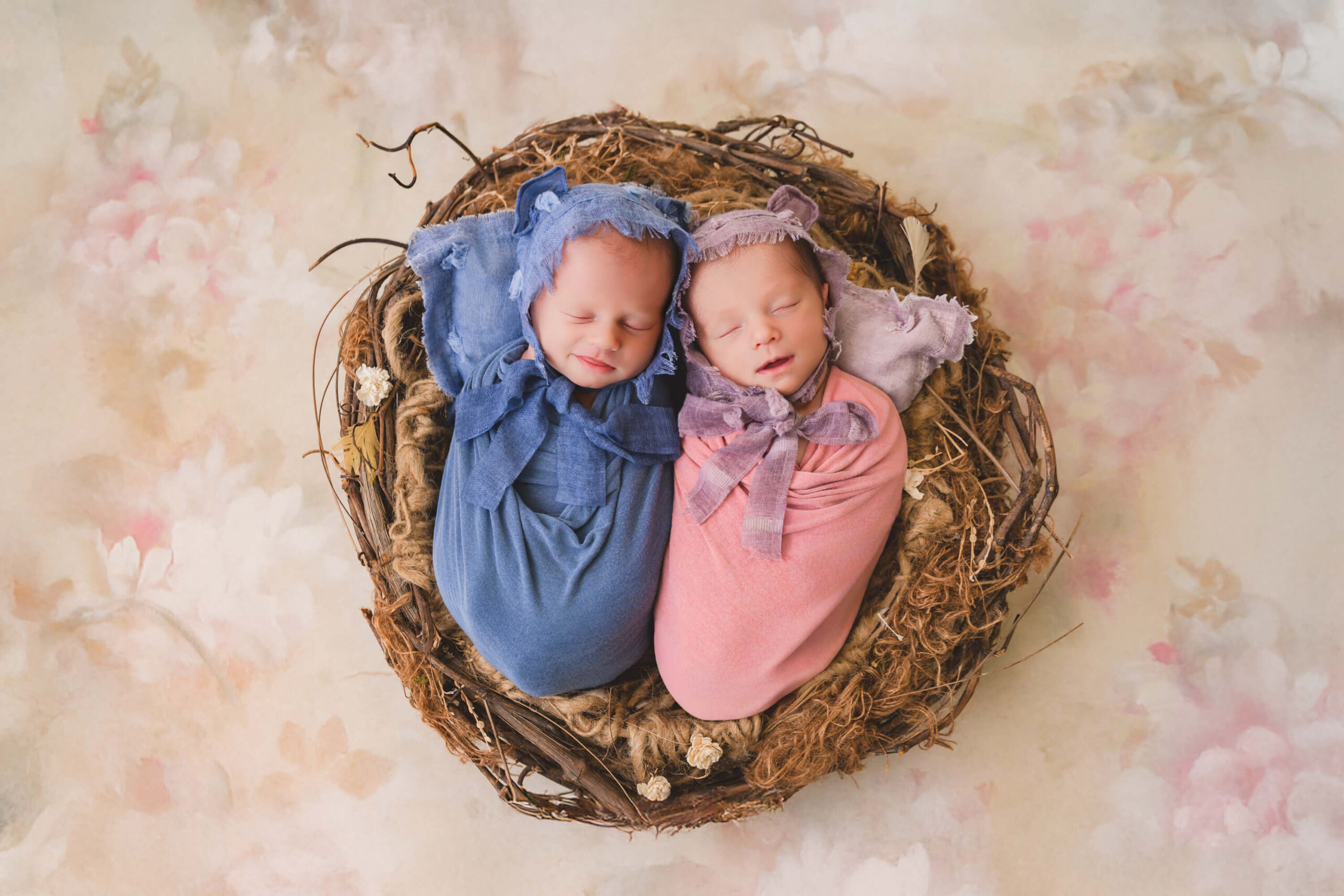 Newborn twin brother and sister sleep peacefully in a bird nest basket in matching pink and blue swaddles and bonnets after finding organic baby clothes in san francisco