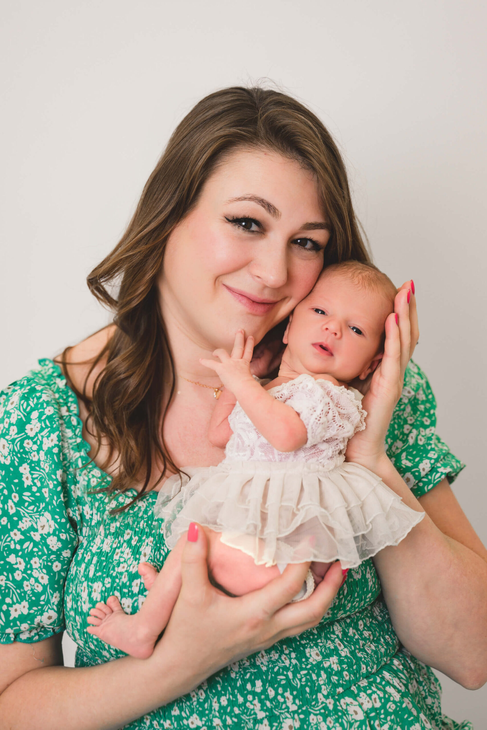 A happy new mom in a green dress cradles her awake newborn daughter in a white gown against her cheek after finding organic baby clothes in san francisco