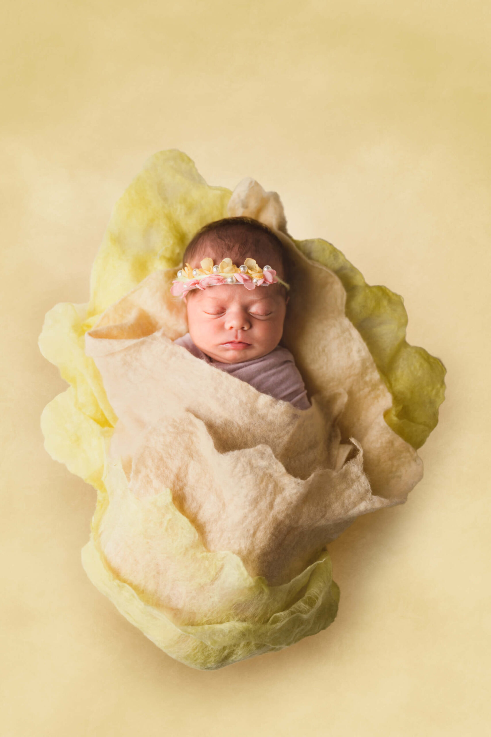 A sleeping newborn girl in a flower and pearl headband in a felt wrap in a studio